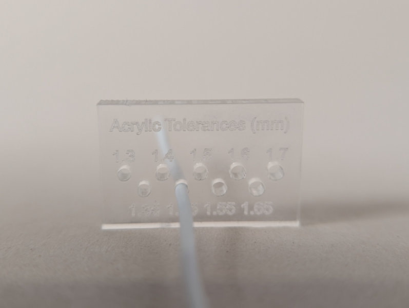transparent rectangle with "acrylic tolerance" and numbers etched into it and small holes. A wire passes through one of the center holes