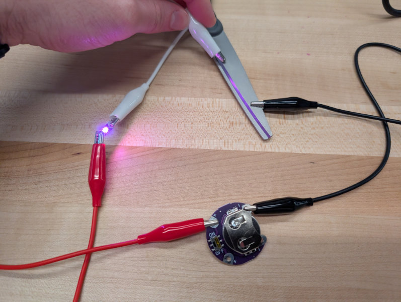 red and black alligator clips connecting battery to pink LED and attaching to metal scissors that close the circuit