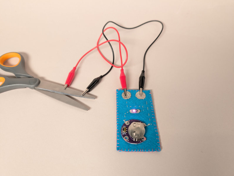 Blue felt rectangle with thread wiring, battery and pink LED, connected to red and black alligator clips that attach to metal scissors