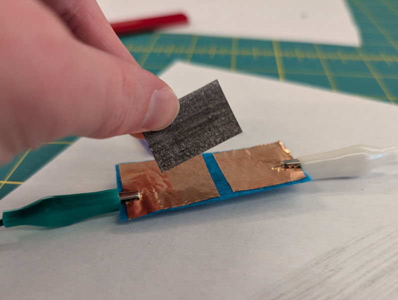 fingers holding graphite-covered paper piece over blue felt rectangle with square copper pads