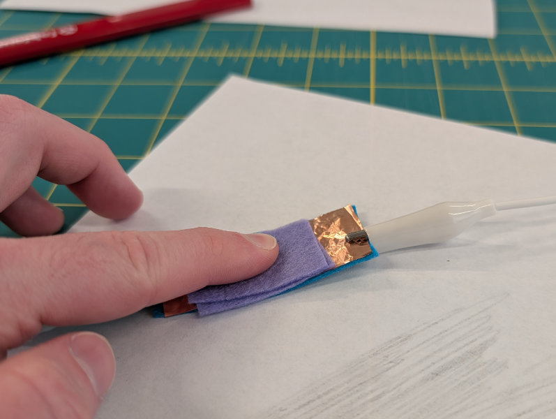 finger pressing down on purple felt over copper pads with alligator clips coming out from the edges