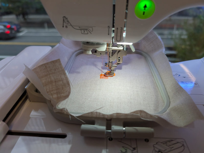 sewing machine closeup running over embroidery hoop with natural linen fabric and orange thread